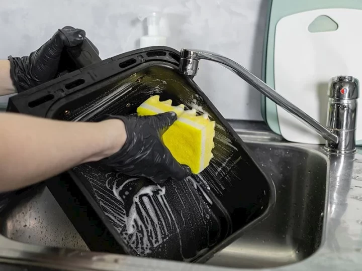 Siyah eldivenle sarı sünger kullanılarak lavaboda airfryer sepeti yıkanıyor