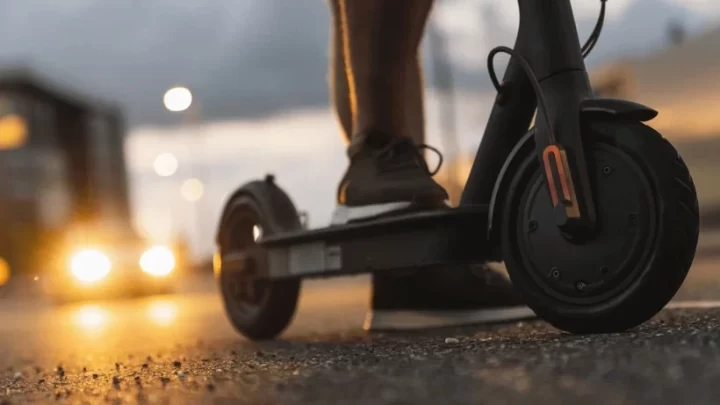 Elektrikli scooter’ın tekerleği ve üzerinde duran bir kişinin ayakları yakın planda görülüyor.
