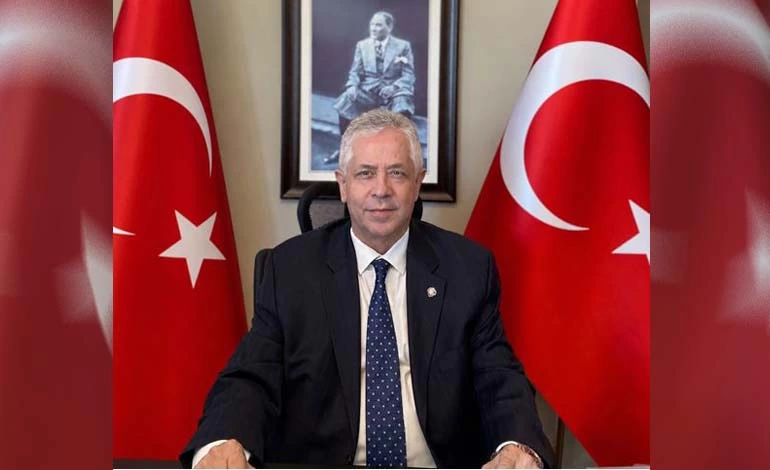 Müjdat Güler, Türk bayrakları ve Atatürk portresi önünde masasında oturuyor.
