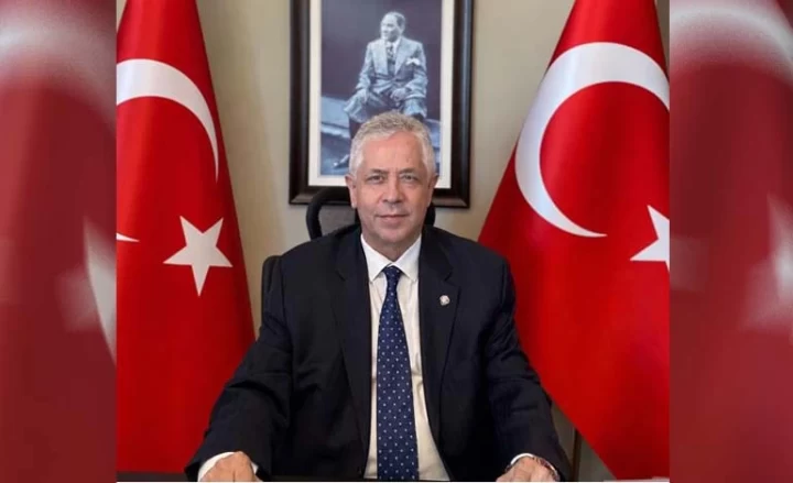 Müjdat Güler, Türk bayrakları ve Atatürk portresi önünde masasında oturuyor.