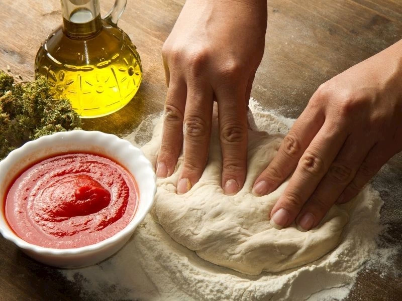 Eller pizza hamurunu unlanmış tezgahta şekillendiriyor; yanında domates sosu ve zeytinyağı var