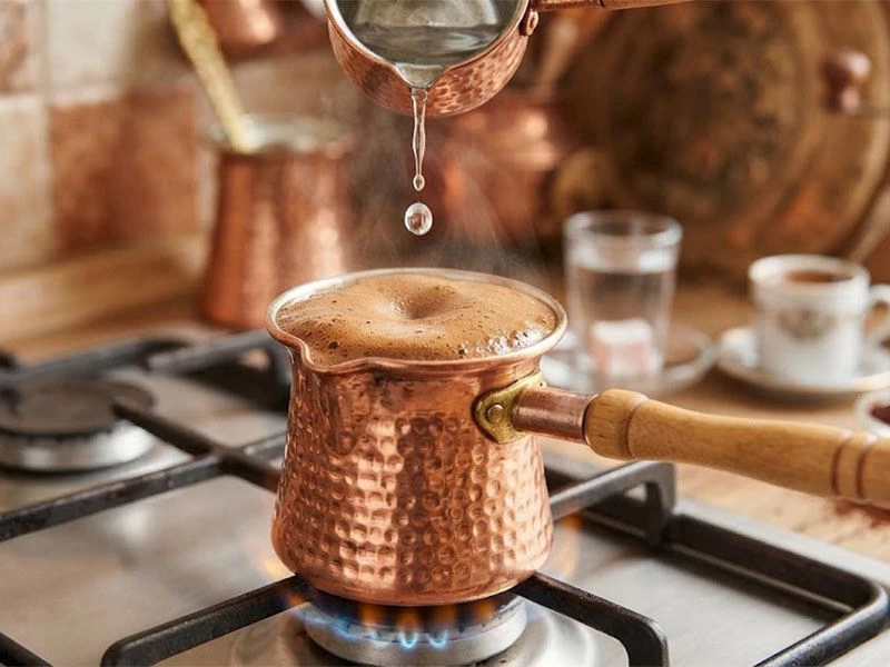 Ocaktaki bakır cezvede köpüklü kahve pişerken üzerine su damlatılıyor